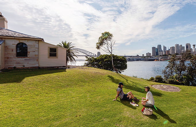 A young woman and man sit on the grass next to a historic building on Goat Island, with view of the harbour and Sydney in the background. Photo: David Finnegan &copy; DPIE