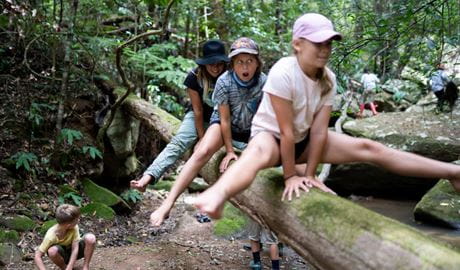 Three children wearing hats leapfrog over a fallen tree branch in the bush. Credit: Blanc Content Photography &copy; Blanc Content Photography