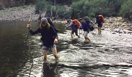 Hikers with backpacks make a river crossing in a wilderness setting. Photo credit: Adam Stewart &copy; The Tops Conference Centre