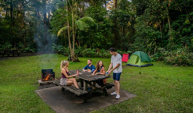 Four people sit at a picnic table beside a campfire at Sheepstation Creek campground, Border Ranges National Park. Photo credit: John Spencer &copy; DPIE