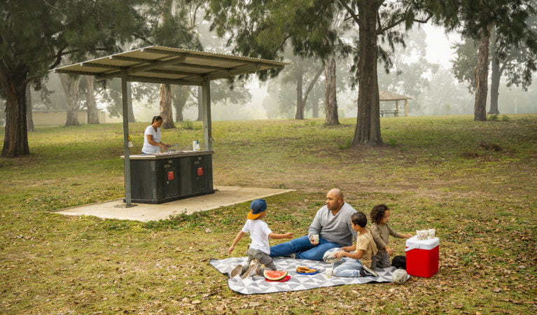 A man and his children sit on a picnic blanket while a woman cooks lunch on the barbecue, at Cattai Farm picnic area in Cattai National Park. Photo: John Spencer/OEH