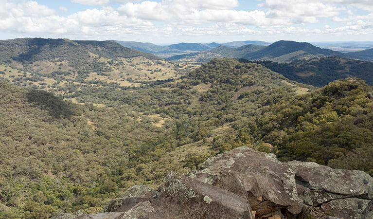 View past a rocky ledge to a rolling landscape dotted with bushland, in Coolah Tops National Park. Photo: Leah Pippos/DPIE