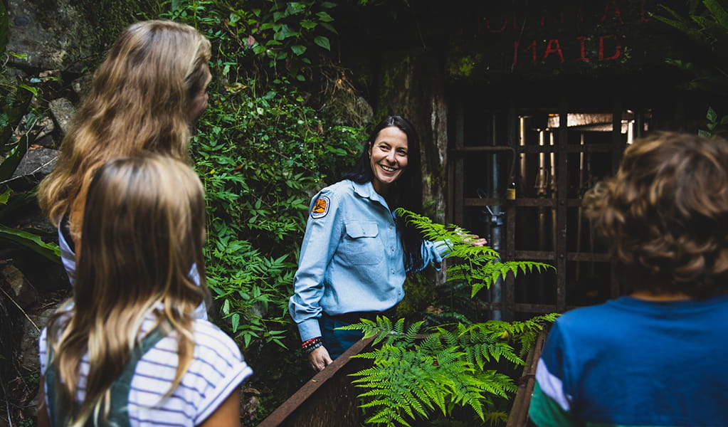 Family enjoying a tour of Mountain Maid gold mine surrounded by ferns and looking through a doorway grate with a ranger. Photo credit: Destination NSW &copy; Destination NSW