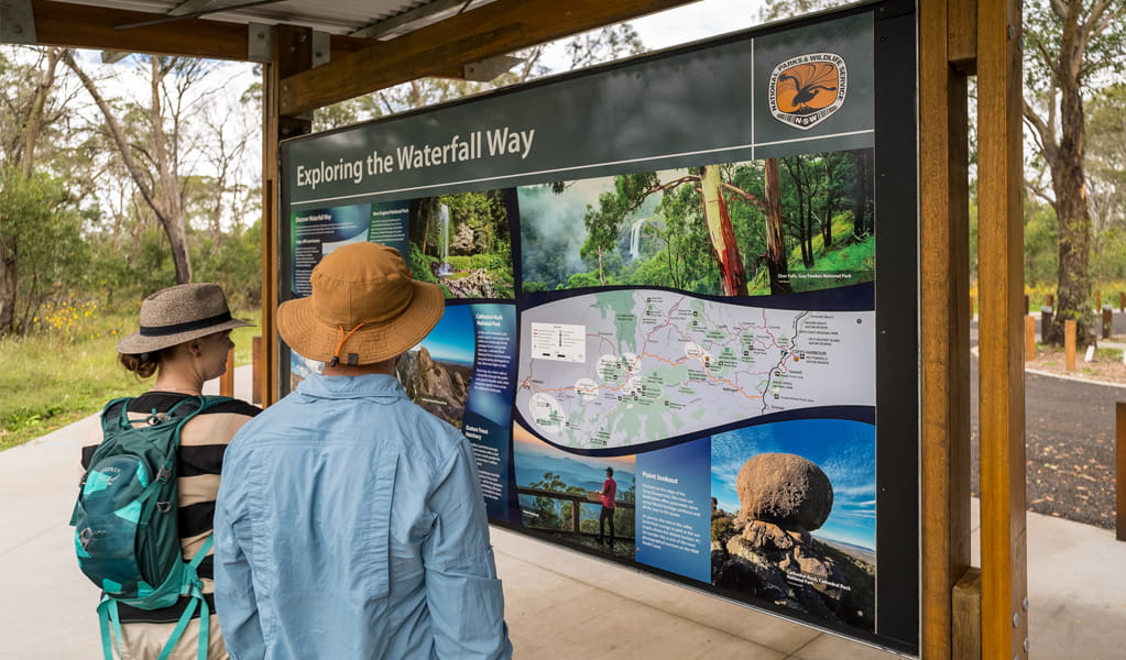 Two visitors looking at a colourful sign about Waterfall Way at Ebor Falls picnic area, Guy Fawkes River National Park. Photo: David Waugh, &copy; DCCEEW