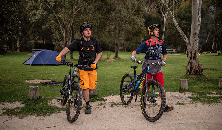 Two mountain bike riders walk past a tent at Ngarigo campground, Kosciuszko National Park. Photo: Robert Mulally/DPIE