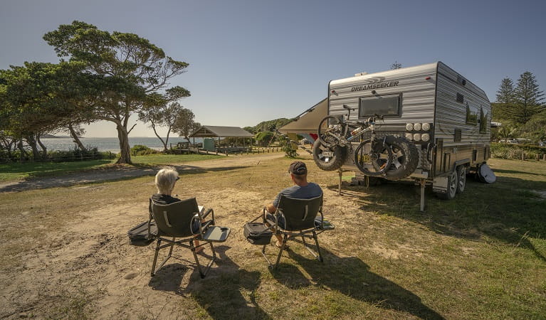2 people sitting down beside their campervan looking out at the ocean at Point Plomer campground, Limeburners Creek National Park. Photo: John Spencer/OEH