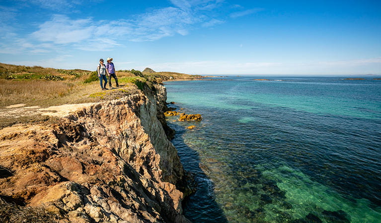 2 visitors look out to the ocean from a cliff top, with coastal heathland in the background. John Spencer &copy; DPIE