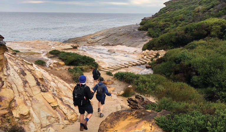 Walkers descending an ochre-coloured sandstone slope before crossing a creek using large stepping stones, with views out over the ocean. Photo: Natasha Webb