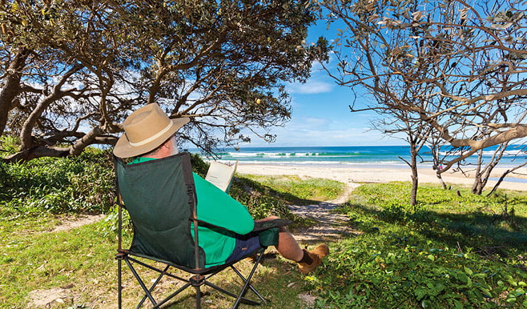 A man reads a book in the shade with a view of the beach in Red Cliff campground, Yuraygir National Park. Photo: Robert Cleary/DPIE