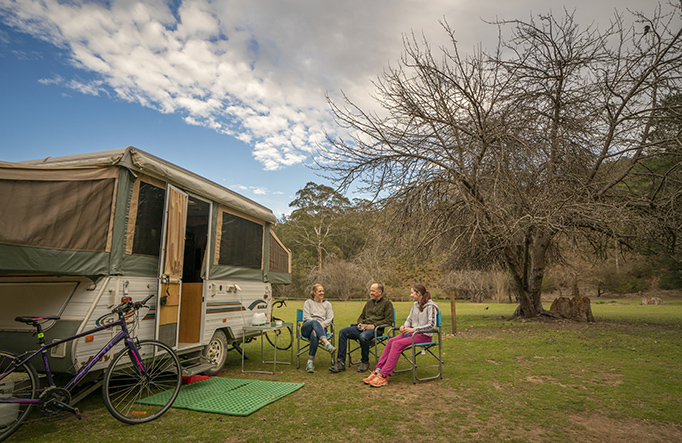 Three people sit on camp chairs beside a caravan at Wombeyan Caves campground. Photo: John Spencer/DPIE