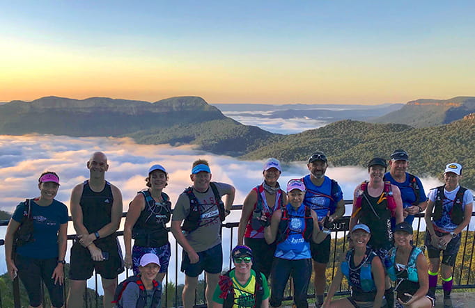 A group of runners stand on a lookout platform in front of a sweeping view of mountains and mist-filled valleys. Photo credit: Tony Williams copy; Blue Mountains Fitness