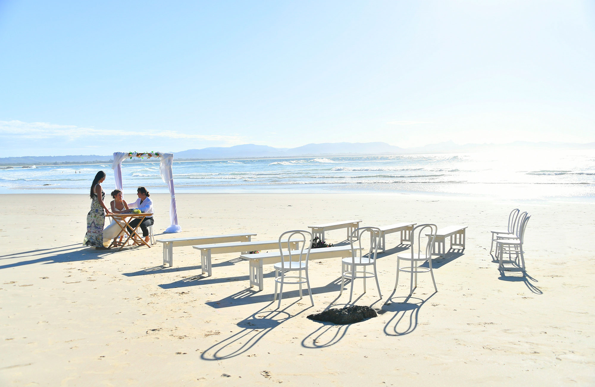 A couple and their celebrant next to a wedding arch with chairs and tables in the foreground on Wategos Beach in Cape Byron State Conservation Area. Photo: Fiora Sacco &copy; DPIE