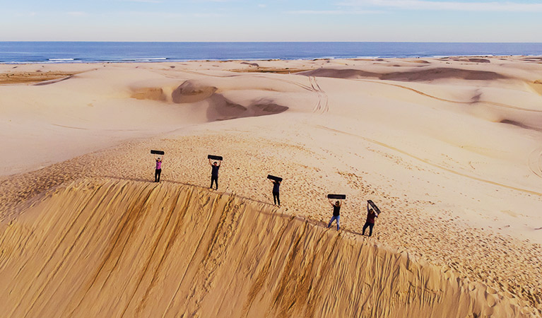 Aerial view of 5 people standing at the top of a massive sand dune, holding their sand boards in the air. Photo &copy; 4WD Tours R Us