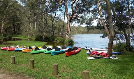 View of 2 brightly-coloured kayaks at the shoreline of Myall Lake, with the sun setting behind trees. Photo credit: Tony Schofield &copy; Adventure Activities Outdoors  