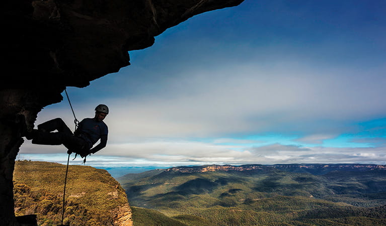 A silhouetted climber descends by rope beneath a cliff overhang, against a backdrop of sky and distant green valleys and mountains. Photo credit: David Hill &copy; Blue Mountains Adventure Company