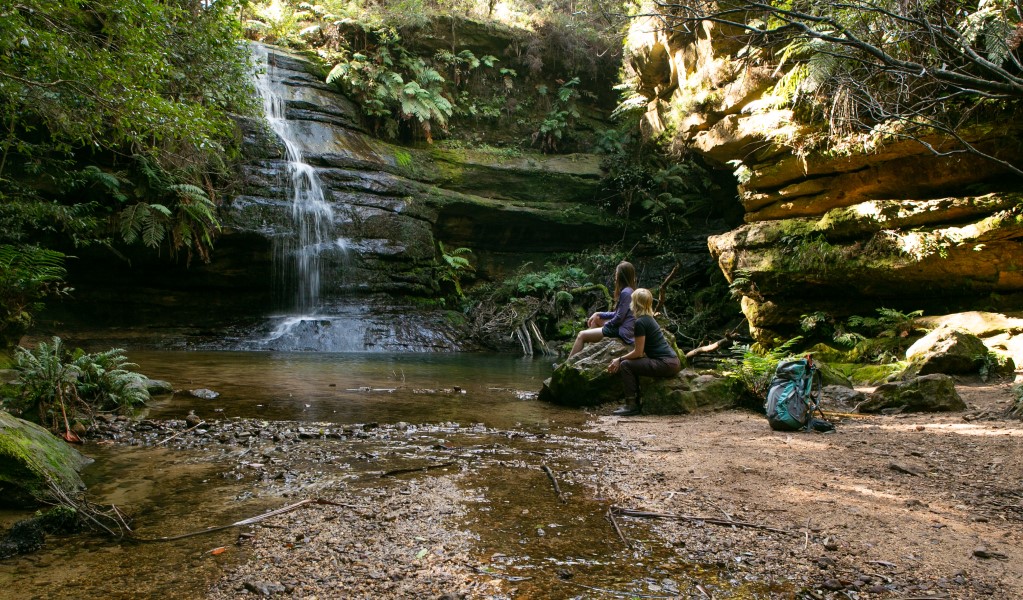Two hikers rest near a waterfall surrounded by mossy rocks and green ferns at Pool of Siloam in Blue Mountains National Park. Photo: Jannice Banks &copy; Blue Mountains Adventure Company