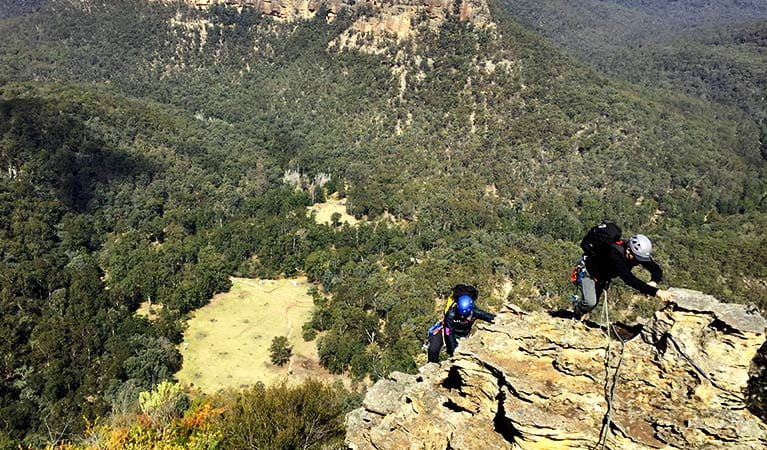 2 climbers wearing helmets climb over a rugged sandstone ledge with forest-clad mountains in the background. Photo credit: Hugh Ward &copy; Blue Mountains Climbing School