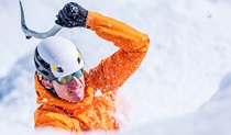 A man wearing an orange jacket and white helmet swings his ice axe in a snow-covered landscape. Photo &copy; Martin Cankov