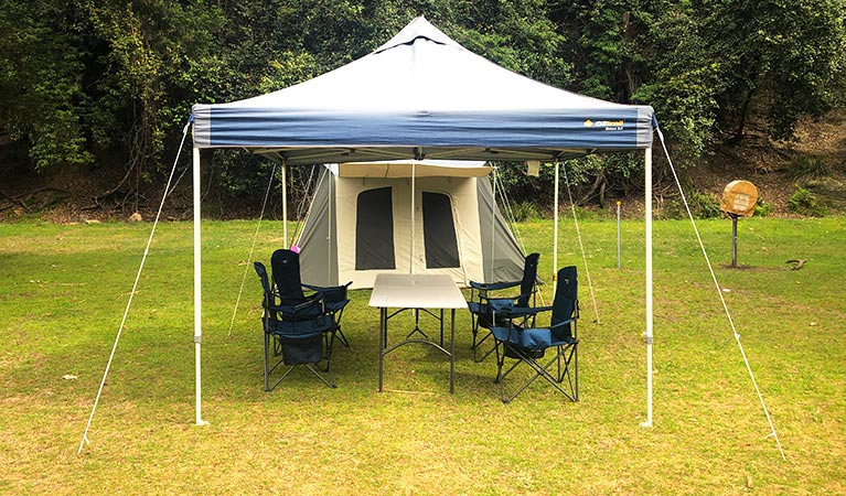 A pitched tent with chairs and a tent canopy set up on a grassy clearing, with bushland in the background. Photo credit: Damien McClellan &copy; EcoTreasures