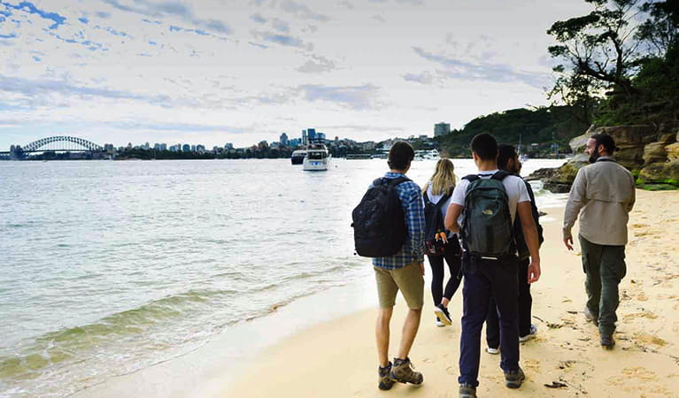 A group treks along a sandy beach with their EcoWalks Tours guide, with the Sydney Harbour Bridge in the distance. Photo © EcoWalks Tours