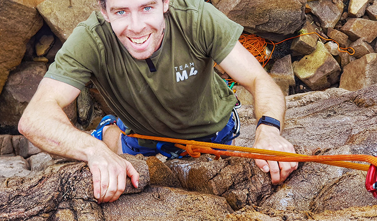 A young man smiles as he grips rock holds and climbs up a sheer cliff whilst roped in. Photo © Nash Hall