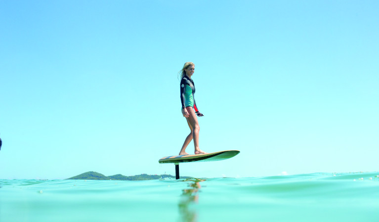 A woman in a wetsuit riding on a e-foil board across water with a blue sky in the background. Photo: Fliteschool Manly &copy; Fliteschool Manly