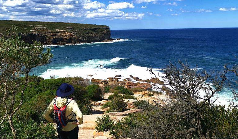 A bushwalker with a day pack looks out over surf breaking on a wild rocky coastline. Photo credit: Sara Freeland © Freeland Hiking Co