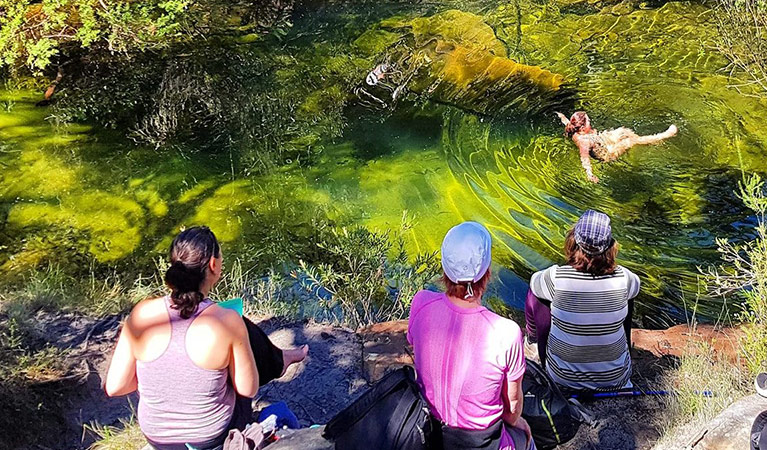A group of 3 women sit along the bank of a natural pool as another women swims. Photo credit: Sophie Orwin © Heart Hum