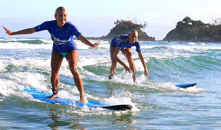 A pair of women in Le'ts Go Surfing rash shirts smile as they ride a small wave near Byron Bay. Photo &copy; Let's Go Surfing