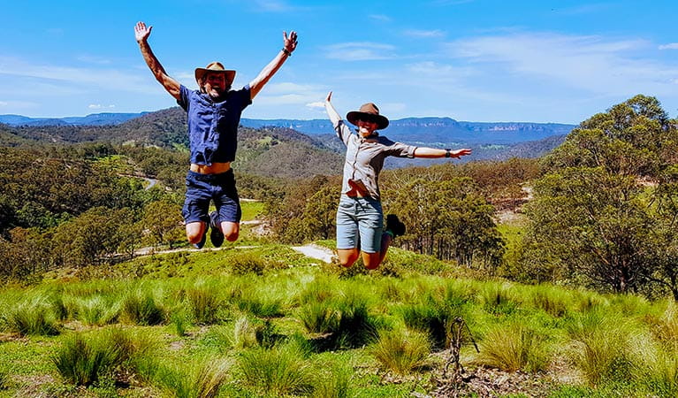 A man and woman leap into the air near a walking track, with cliffs and valleys of the Blue Mountains in the background. Photo &copy; Life's An Adventure.