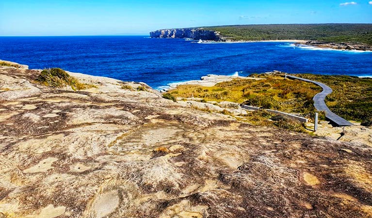 View of coastal rock formations with a winding boardwalk, ocean bay and coastal cliffs in the background. Photo credit: Kerry Martin © Lost and Found Adventures