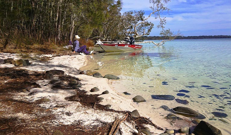 A woman seated on a lakeshore near a moored motorboat looks out over clear waters to a distant shore. Photo credit: Peter Smee © Manning Valley Adventures