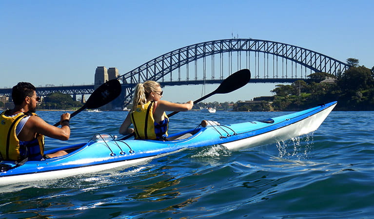 A man and woman paddle a double kayak on Sydney Harbour with the Sydney Harbour Bridge in the background. Photo credit: Matt Bezzina &copy; Paddle Pirates 