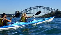 A man and woman paddle a double kayak on Sydney Harbour with the Sydney Harbour Bridge in the background. Photo credit: Matt Bezzina &copy; Paddle Pirates 