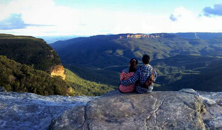 A couple seated on a rocky ledge gaze out over a vista for forest-clad mountains and valleys In Blue Mountains National Park. Photo credit: Dave Hadari &copy; Oz Trails 