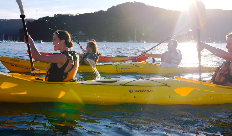 View of a group of smiling kayakers paddling across still waters against a burst of sunlight. Photo © Spencer Frost