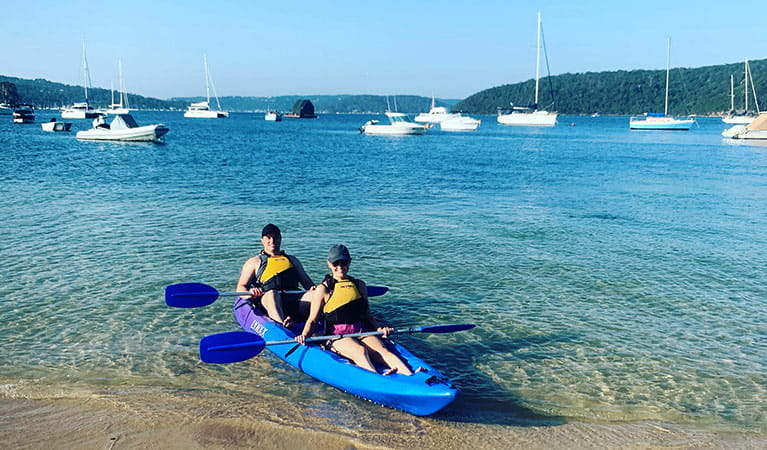 View of 2 people in a double kayak at the shore of a sandy bay, with boats in the background. Photo credit: Greg Moran &copy; Pittwater Kayak Tours
