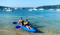 View of 2 people in a double kayak at the shore of a sandy bay, with boats in the background. Photo credit: Greg Moran &copy; Pittwater Kayak Tours