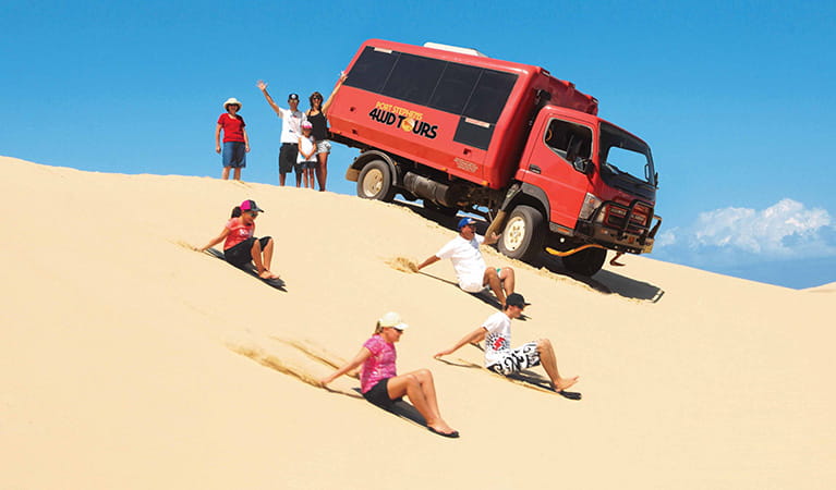 A group enjoys sand boarding on the dunes alongside a red Port Stephens 4WD Tours vehicle. Photo credit: Ray Alley &copy; Port Stephens 4WD Tours