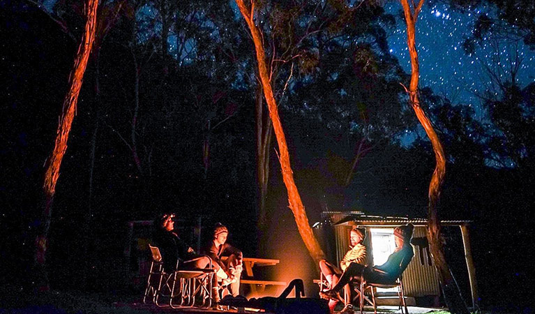 A group of people sit around a glowing campfire next to a hut beneath starry skies. Photo credit: Benny Littlejohn  &copy; Primal Adventures