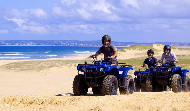 3 people wearing helmets ride blue quad bikes along coastal dunes. Photo &copy; Quad Bike King