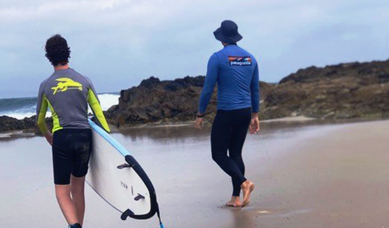 A surfer and his Rusty Miller Surf instructor walk across a beach toward breaking waves. Photo &copy; Rusty Miller Surf