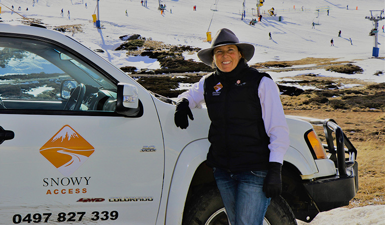 A smiling woman leans on a white vehicle emblazoned with the Snowy Access logo, with ski fields in the background. Photo © Claire Rogerson