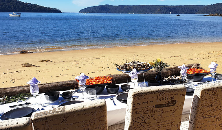 View of chairs and table set for fine dining, against a backdrop of a sandy beach and sheltered bay. Photo &copy; Andriana Papandreau