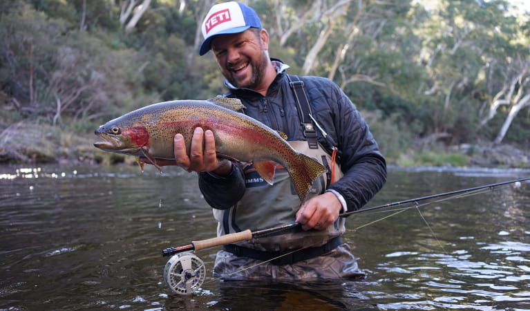 Man holding a rainbow trout and fishing rod wading in a river. Photo: M. Tripet/The Fly Program