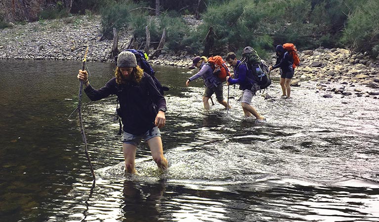 Hikers with backpacks make a river crossing in a wilderness setting. Photo credit: Adam Stewart &copy; The Tops Conference Centre