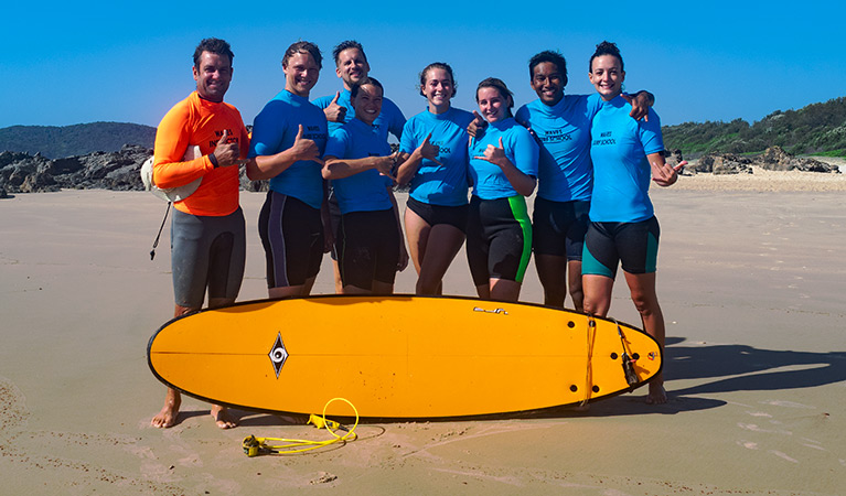 A group of surfers and their instructor pose with a surfboard on the beach. Photo credit: Annemieke Yperlaan © Waves Campground