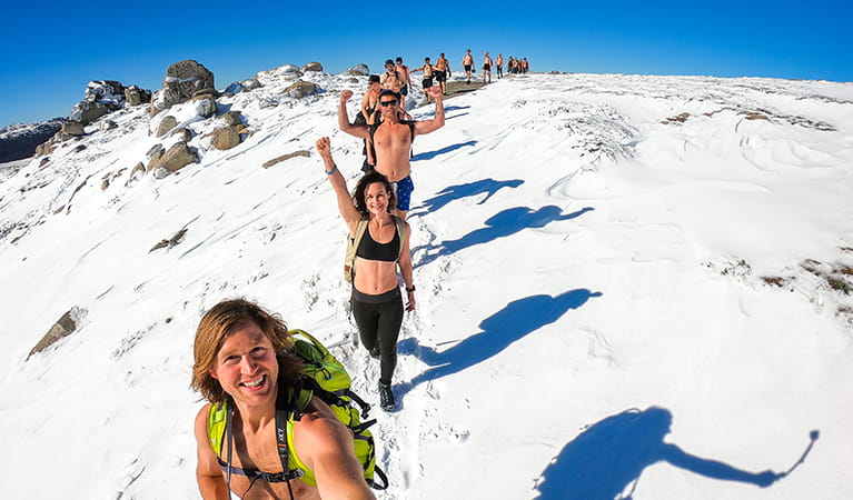 Trekkers, some dressed only in shorts, hike in a long line across a snowy alpine landscape in Kosciuszko National Park. Photo &copy; Majell Backhausen