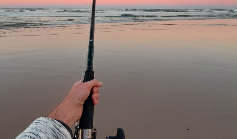 View of an angler's arm with a fishing rod and reel on a pristine sandy beach at twilight. Photo credit: Tim Williams &copy; Wilderness Adventures
