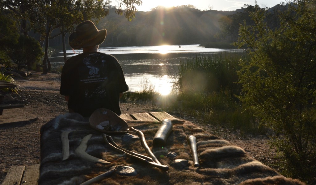 A Wirimbili Cultural Experience guide with a display of traditional Aboriginal tools overlooking serene river waters surrounded by bushland at Ganguddy-Dunns Swamp in Wollemi National Park. Photo: Cindy Budai &copy; Wirimbili Cultural Experience